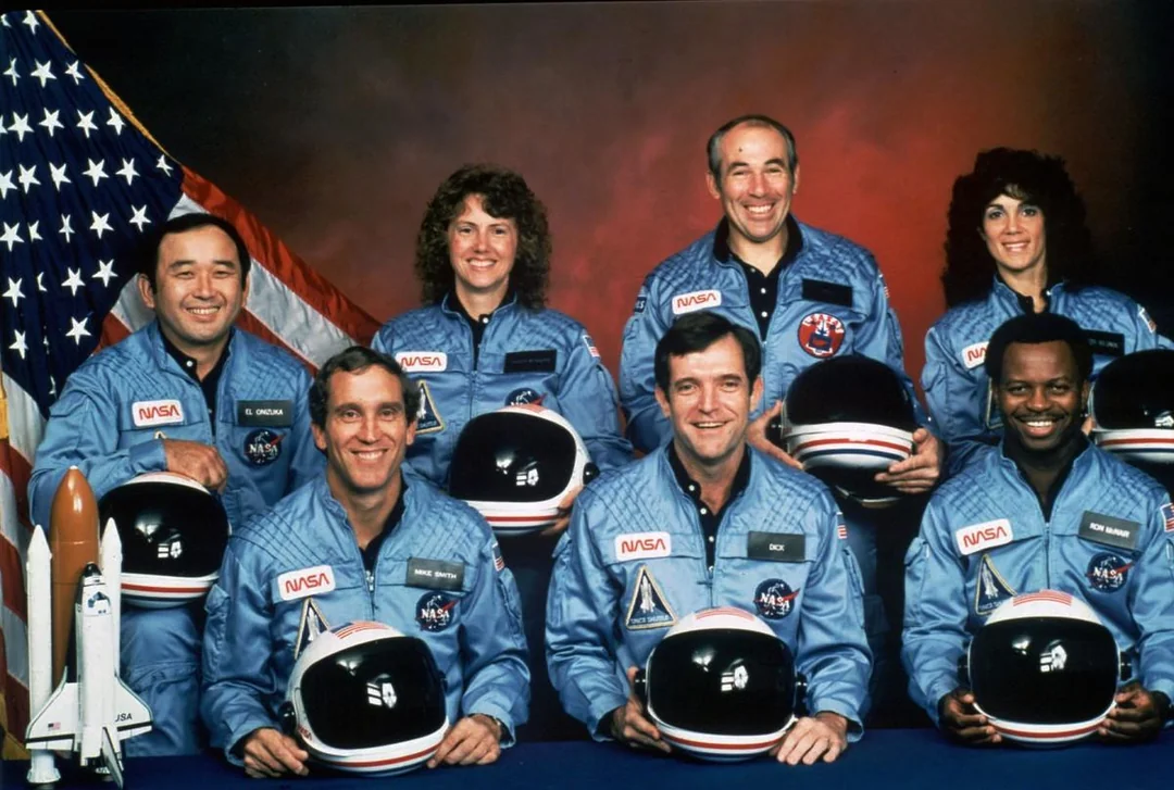 Seven astronauts in blue NASA uniforms pose, smiling, with helmets in hand in front of a U.S. flag and a model of a space shuttle. Four people stand in the back row, and three sit in the front row.