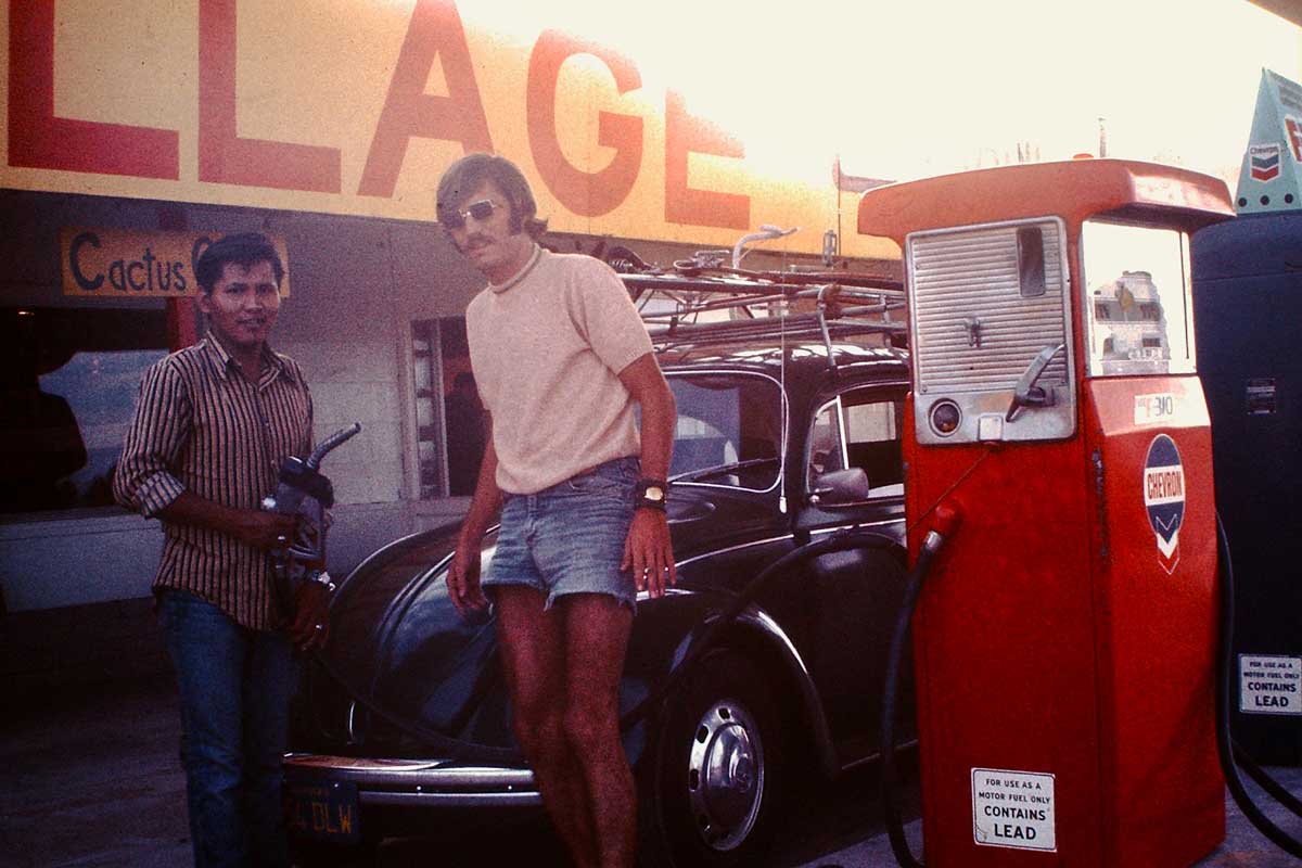 Two men stand by a red gas pump, refueling a black Volkswagen Beetle at an old-fashioned gas station with a yellow and orange sign in the background.
