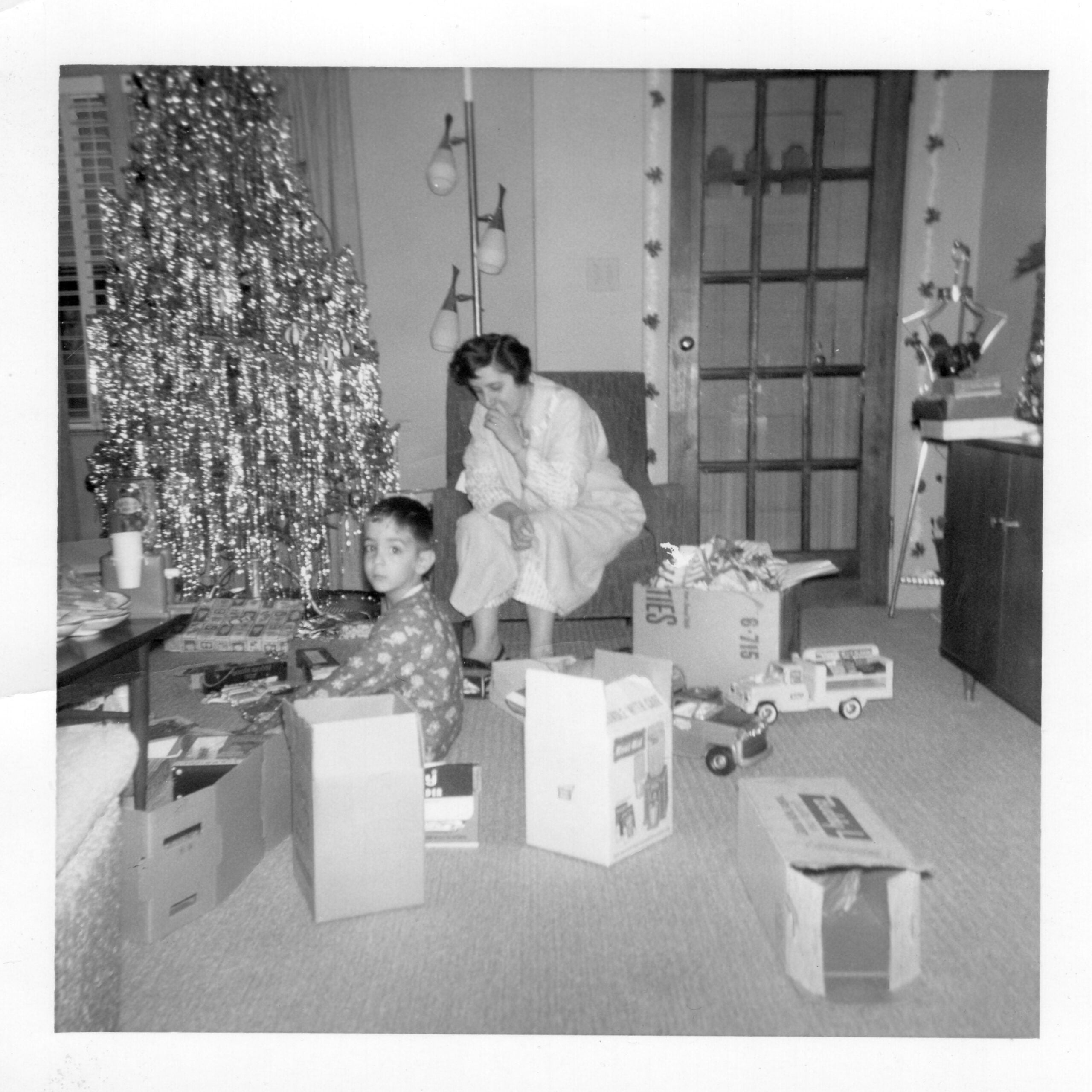 A young boy sits among boxes and toys near a tinsel-covered Christmas tree, while a woman in a robe sits behind him, watching. The room is decorated festively, with presents and wrapping paper scattered around.