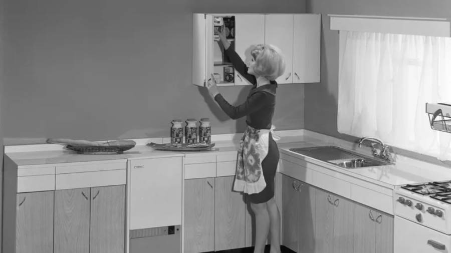 A woman in a dress and apron reaches up to open a kitchen cabinet in a vintage-style kitchen, with jars and a loaf of bread on the counter.
