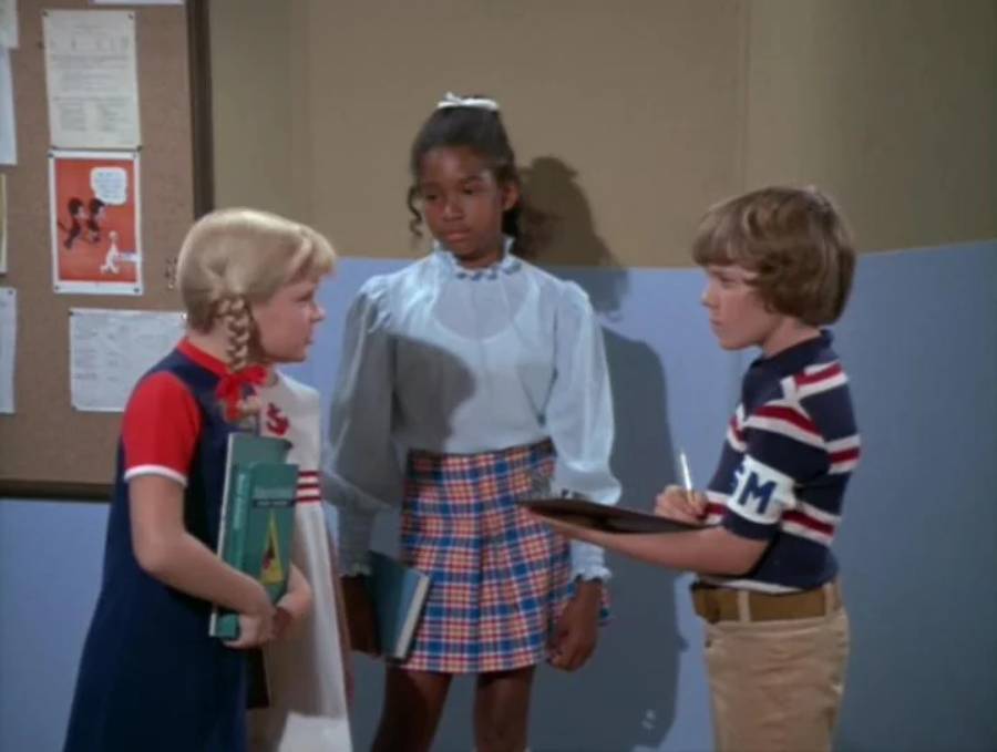 Three children stand indoors near a bulletin board. One girl with braids holds books, another girl in a light blouse and plaid skirt stands behind, and a boy in a striped shirt writes on a clipboard.