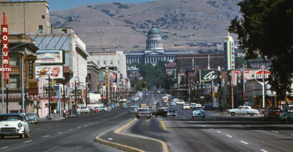 A wide street in a city with vintage cars, lined with shops, hotels, and neon signs. In the background, a domed government building sits at the base of rolling hills under a clear sky.