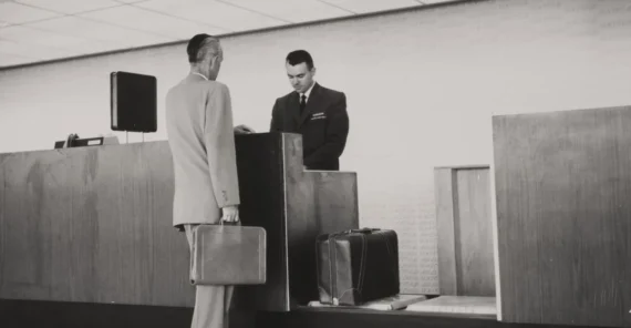 A man in a suit stands at an airline check-in counter with a briefcase, speaking to an airline agent. Two suitcases sit on the counter. The area is labeled "Tickets & Baggage." The photo is in black and white.