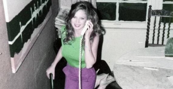 A woman with long hair, wearing a green top and purple pants, smiles while talking on a corded phone in a room with a painting, a round table, and a window.