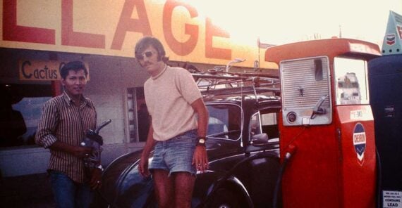 Two men stand by a black Volkswagen Beetle at a vintage gas station. One holds a gas nozzle near an old red pump with a "Contains Lead" sign. The station sign and building are visible in the background.