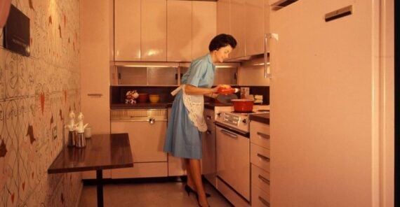 A woman in a blue dress and apron cooks at a stove in a vintage kitchen with pink cabinets, a wall-mounted table, and retro decor.