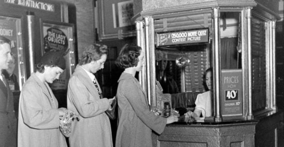 Three women stand in line at a ticket counter, handing money to a cashier behind glass. A man stands behind them, also waiting. The booth displays signs about movie ticket prices and a movie quiz contest.