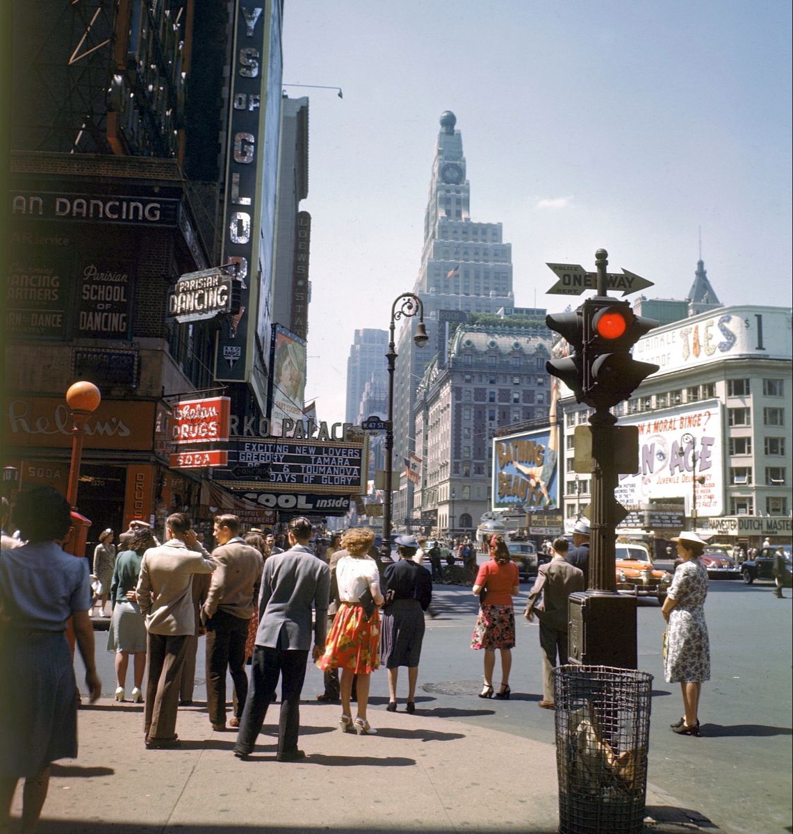 A busy city street scene from the 1940s or 1950s shows people waiting to cross, vintage cars, colorful signs for dancing and drugs, and tall buildings under a clear blue sky.