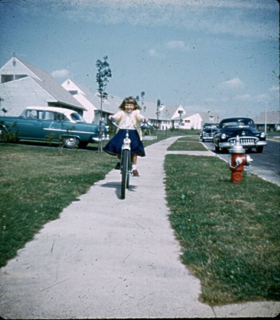 A young girl rides a bicycle on a suburban sidewalk lined with grass, classic cars, and houses under a blue sky with clouds. A red fire hydrant is visible on the right side of the sidewalk.