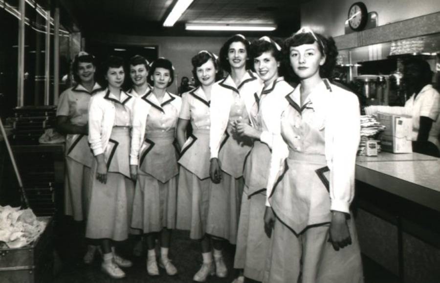 Eight women in matching uniforms stand in a diner or café, smiling at the camera. The uniforms include dresses with aprons and collars. The setting appears to be mid-20th century, with counter and kitchen equipment visible.