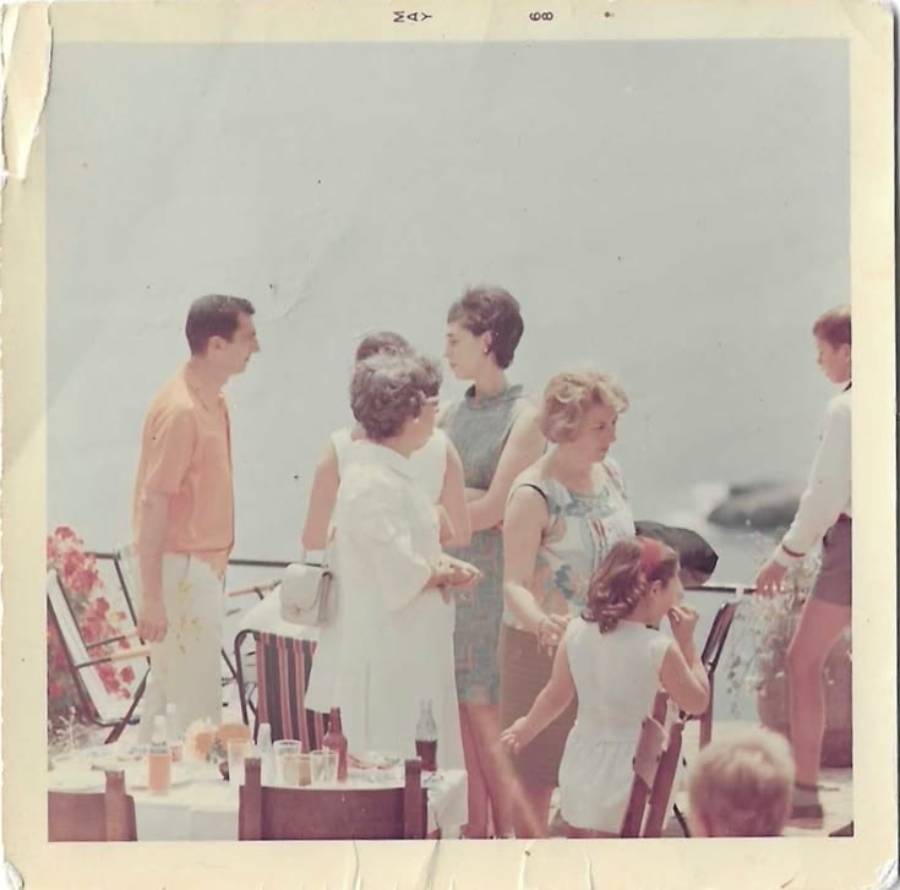 A group of people, mostly women in summer clothes, stand and chat outdoors near a table with drinks; water and a blurred boat are visible in the background.