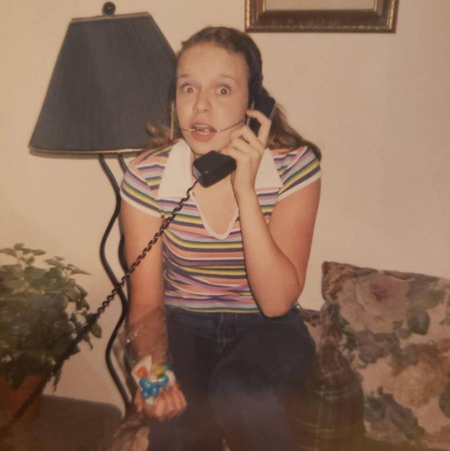 A surprised young woman sits on a couch, holding a corded phone to her ear and a pacifier in her hand. She wears a striped shirt and jeans. A lamp, framed picture, and plant are visible in the background.