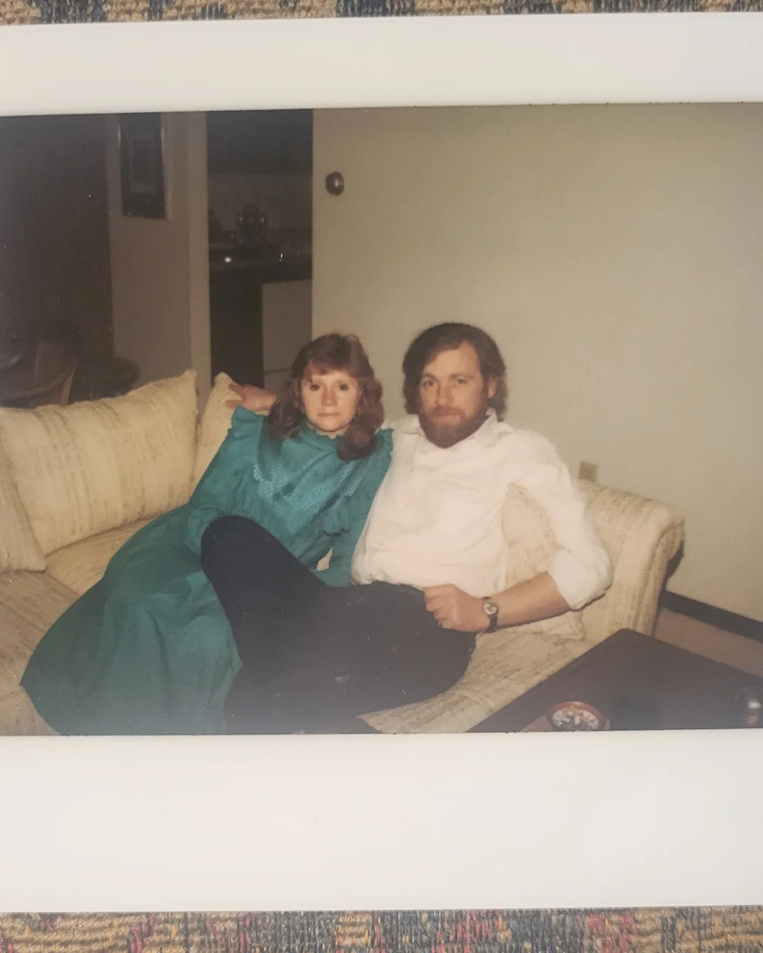 A woman in a green dress and a man in a white shirt sit closely together on a beige couch in a living room, looking at the camera.