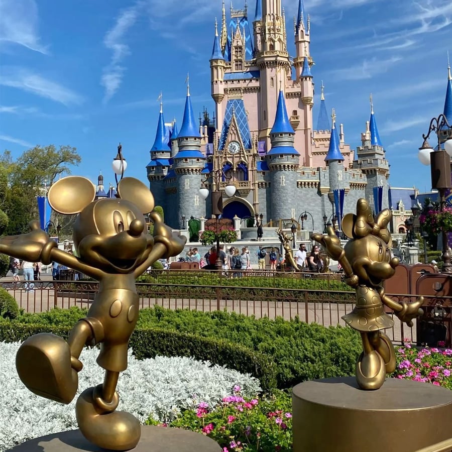 Bronze statues of Mickey and Minnie Mouse stand in front of a pink and blue castle at a Disney theme park, surrounded by green bushes and colorful flowers. Visitors walk in the background under a sunny sky.