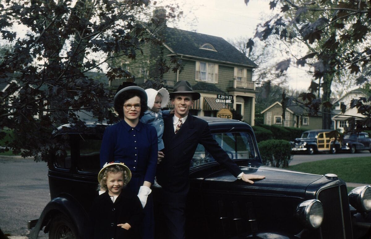 A family of four, dressed in vintage clothing, poses in front of a classic black car on a suburban street with houses and trees in the background.