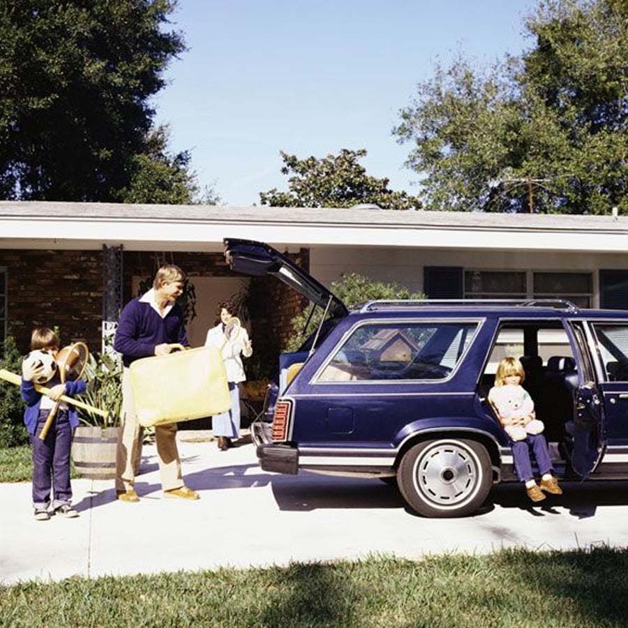 A man loads luggage into a blue station wagon while two children and a woman stand nearby outside a house, preparing for a trip.