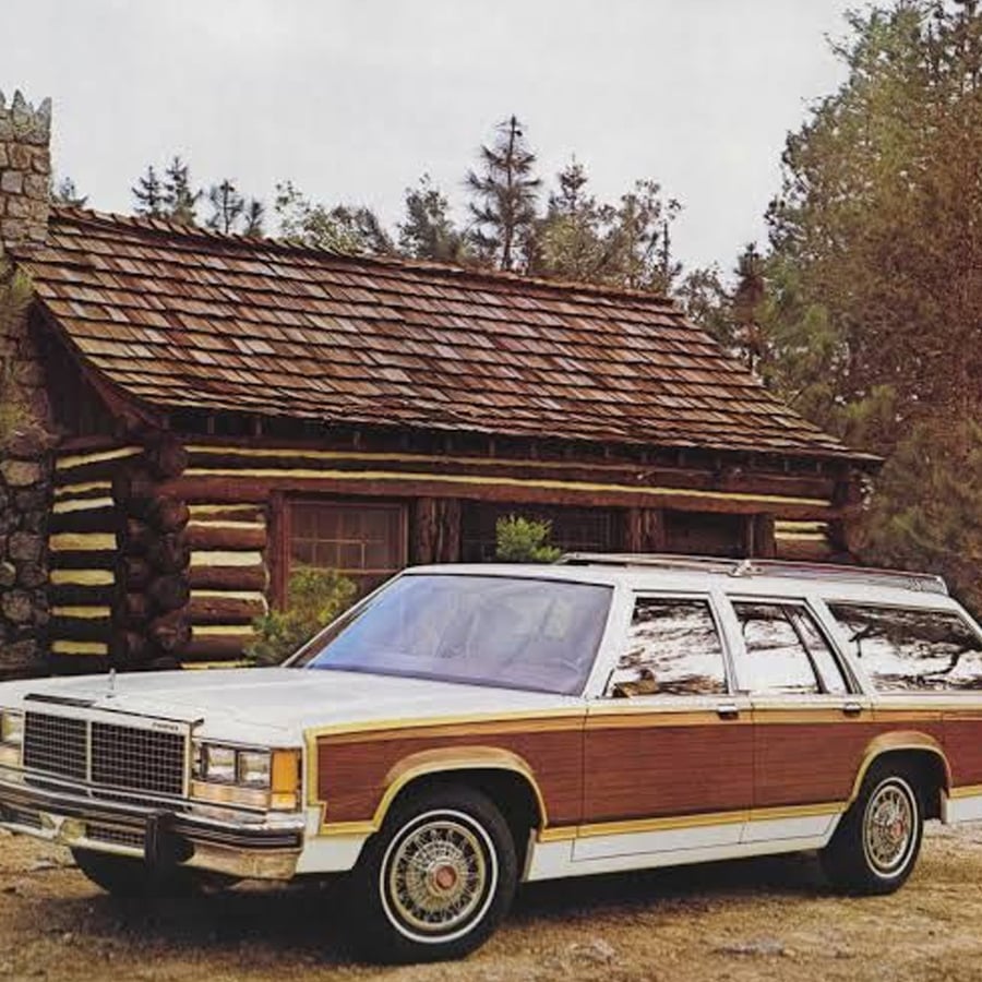 A vintage white station wagon with wood paneling is parked in front of a rustic log cabin surrounded by trees and greenery.