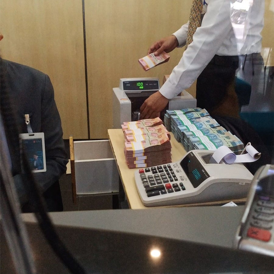 A person counts stacks of banknotes with a counting machine at a counter. There are bundles of cash, a calculator, and a receipt on the desk. Another person sits nearby, partially visible.