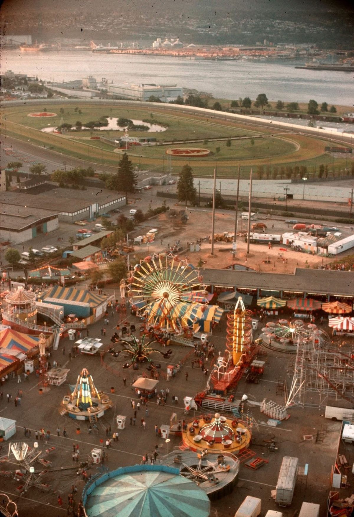 Aerial view of a lively fairground with colorful carnival rides, booths, and crowds of people; a racetrack, river, and cityscape are visible in the background.