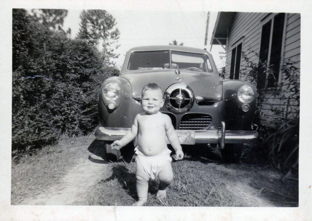 A baby in a diaper stands and smiles in front of an old car parked beside a house, with bushes and trees in the background.