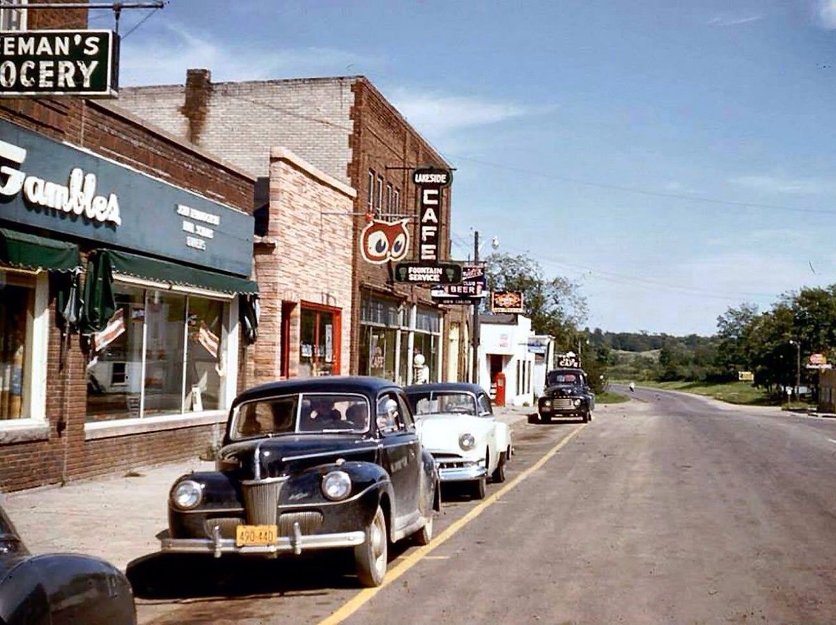 A 1950s small-town street scene with vintage cars parked along a road. Storefronts include "Freeman's Grocery," "Gambles," and a diner with a large "Cafe" sign featuring an owl. Trees and countryside are visible in the distance.