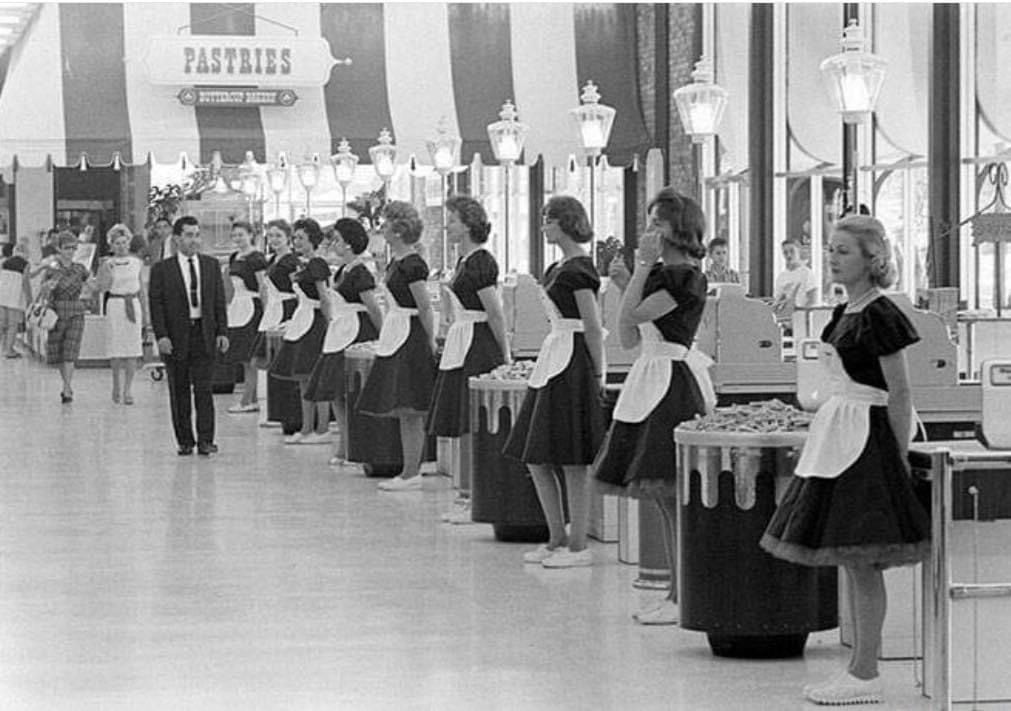 Black and white photo of women in matching dresses and white aprons standing beside checkout counters in a mid-century supermarket, with shoppers and a "Pastries" sign in the background.