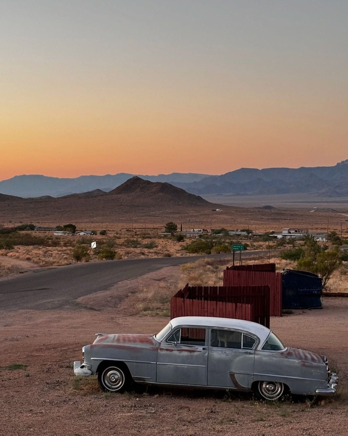 A vintage car with faded paint is parked on dry ground near a road in a desert landscape at sunset, with mountains and sparse buildings in the background.