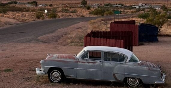 A rusty vintage car is parked on dry grass beside a roadside, with a red fence and dumpster nearby. Desert landscape, mountains, and a soft orange sunset fill the background.
