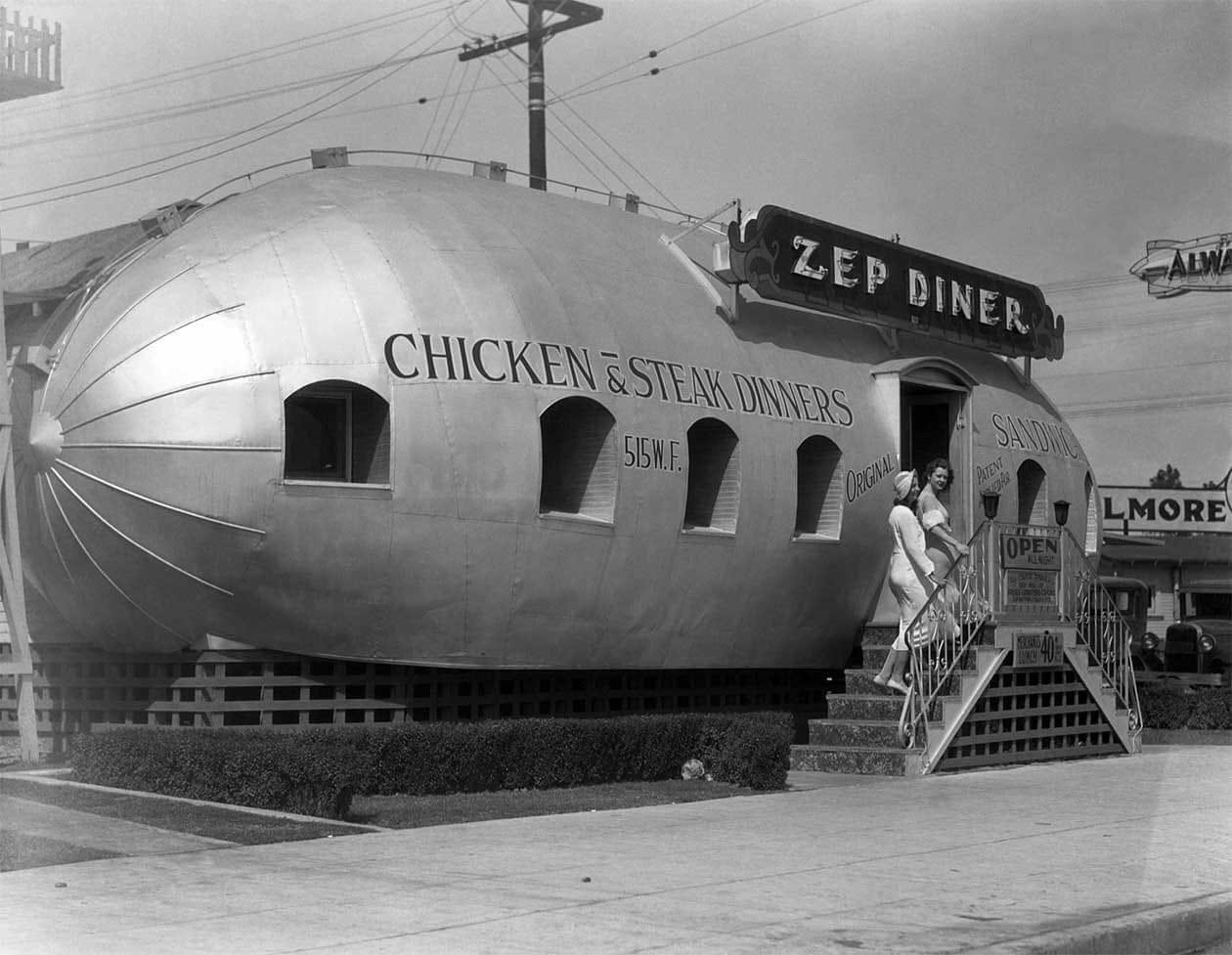 Black-and-white photo of a zeppelin-shaped diner with "ZEP DINER" sign. Two women in white uniforms stand on steps. Text on diner advertises chicken, steak, and sandwiches. "OPEN" sign is on the door.