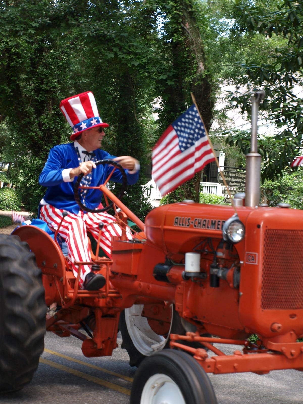 A person dressed in a patriotic Uncle Sam costume drives an orange Allis-Chalmers tractor decorated with an American flag, surrounded by greenery and trees.