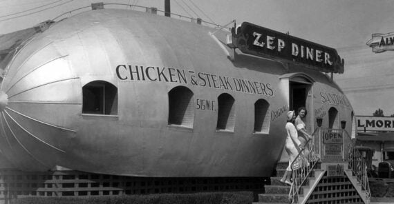 A diner shaped like a zeppelin with "Zep Diner" sign and "Chicken & Steak Dinners" painted on the side. Two people in white uniforms stand by the entrance stairs. 1930s street scene in black and white.