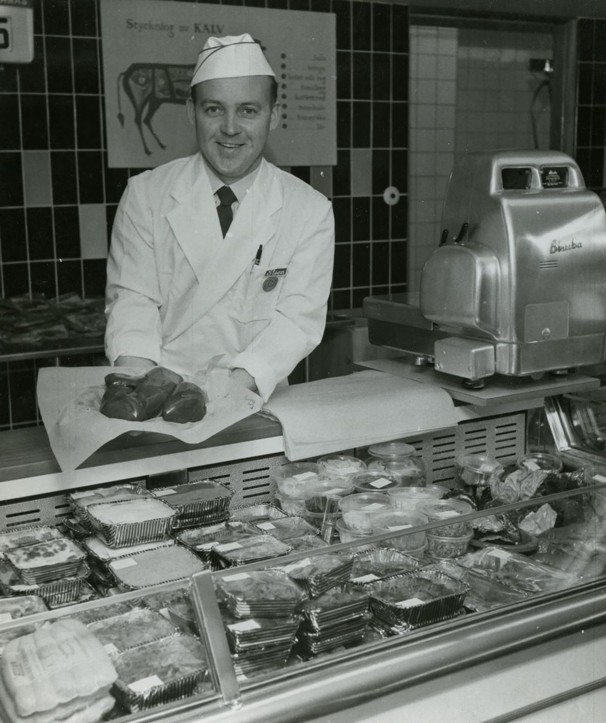 A butcher in a white uniform and hat stands behind a meat counter, holding cuts of meat on paper. Packaged meats and meals are displayed in front, with a meat slicer and a diagram of a cow in the background.