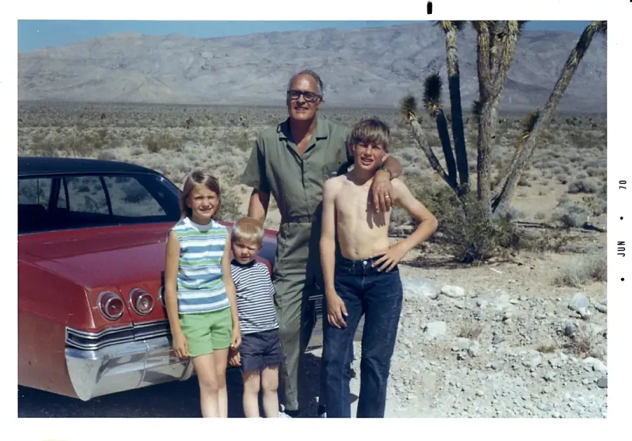 A man stands in the desert with three children near a red vintage car. The group smiles at the camera, with mountains and Joshua trees in the background. The photo has a faded, retro look.