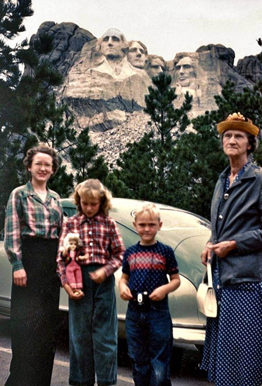 A family of four poses in front of Mount Rushmore, with the famous carved faces visible above trees. Two women and two children stand by an old car; one girl holds a doll, and an elderly woman wears a yellow hat.