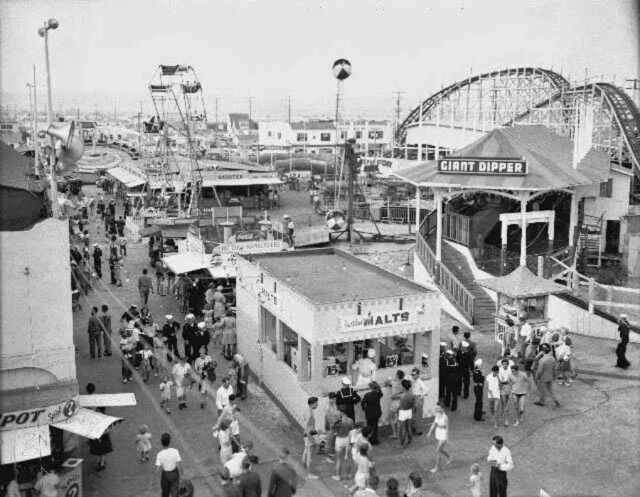 Black-and-white photo of a busy amusement park with crowds walking, food stands, a Ferris wheel, and a roller coaster labeled "Giant Dipper" in the background. People are enjoying various attractions on a sunny day.