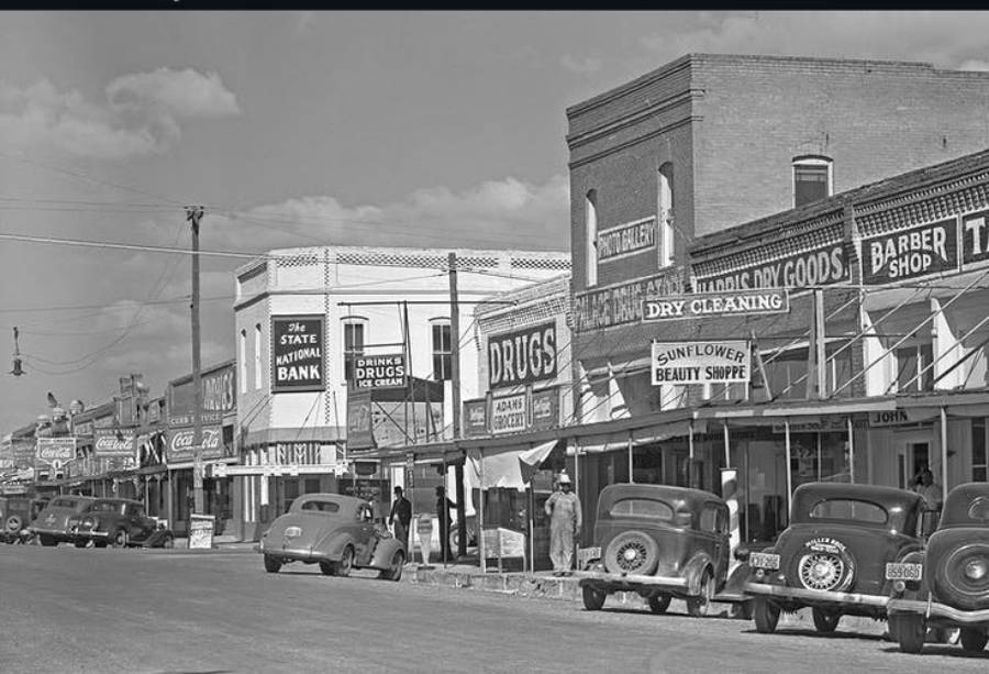 Black-and-white photo of a 1940s small town street with parked vintage cars, people walking, and storefronts including a bank, drugstore, beauty shop, dry cleaner, and barber shop.