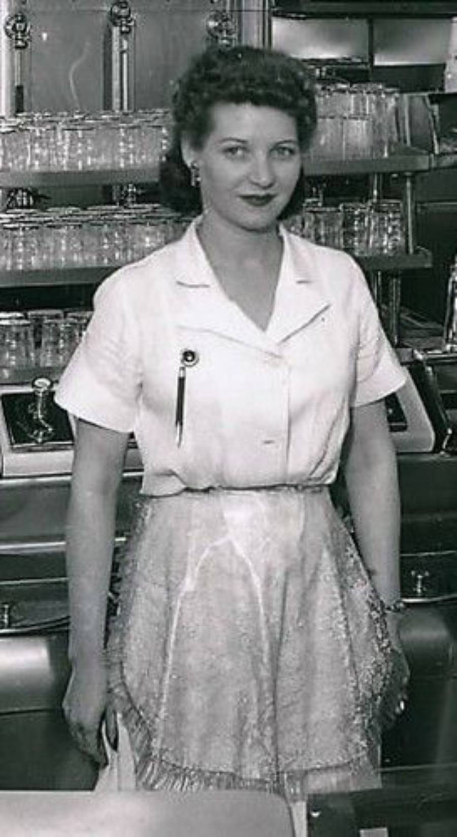 A woman in a white uniform and apron stands in front of a counter with glassware, likely in a diner or restaurant, posing and smiling slightly at the camera.