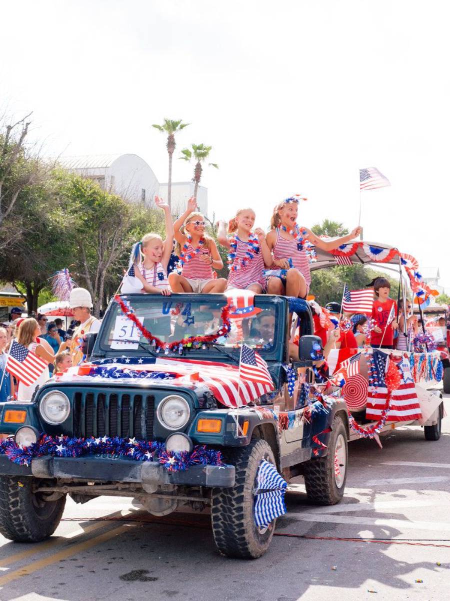 A jeep decorated with American flags and red, white, and blue streamers carries children in festive outfits during a patriotic parade, with a float following behind.