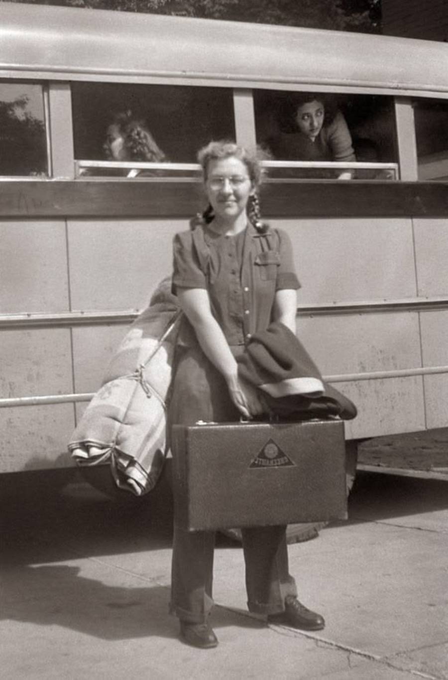 A young woman stands in front of a bus, holding a suitcase and a coat. Two people look out from the bus windows behind her. The photo appears to be from an earlier era, possibly the mid-20th century.