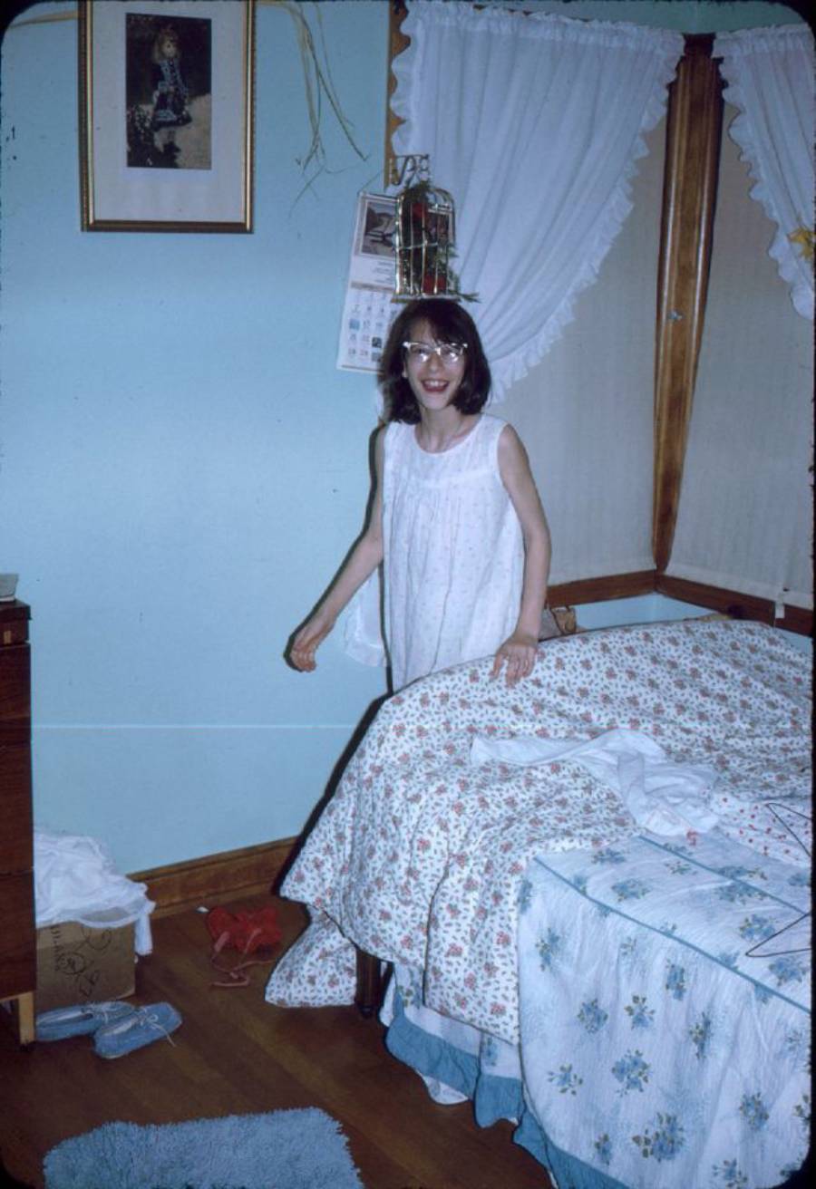 A young girl in glasses and a white nightgown stands by a bed with a floral quilt in a cozy bedroom with blue walls, wooden floors, and curtained windows. Shoes and a cardboard box are on the floor.