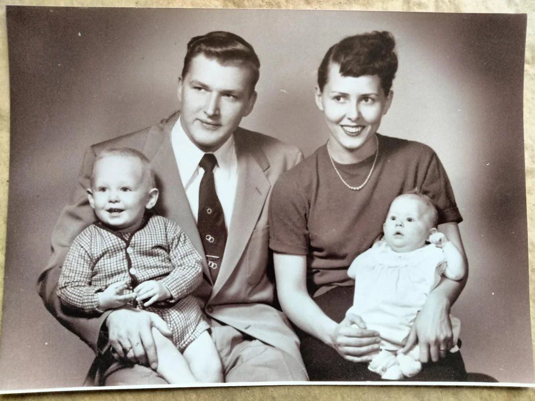 A black and white photo of a smiling man in a suit and a woman in a blouse, each holding a baby. The man holds a toddler in a checked outfit; the woman holds an infant in a light dress.