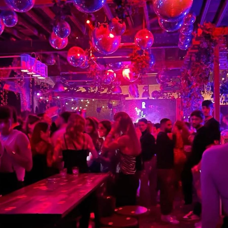 A crowded nightclub with people dancing under pink and purple lights, shiny disco balls hanging from the ceiling, and lush greenery decorating the space.