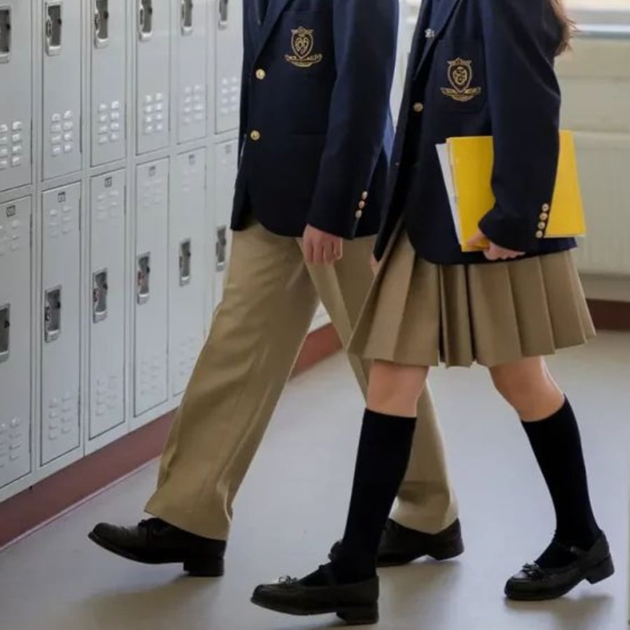 Two students in school uniforms walk past lockers. One wears khaki pants, the other wears a khaki skirt and holds a yellow notebook. Both wear navy blazers and black shoes.