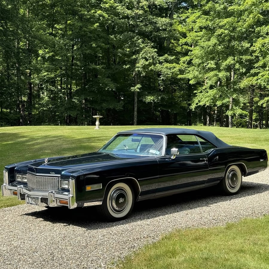 A classic black Cadillac Eldorado convertible is parked on a gravel driveway, surrounded by lush green grass and tall trees in the background.