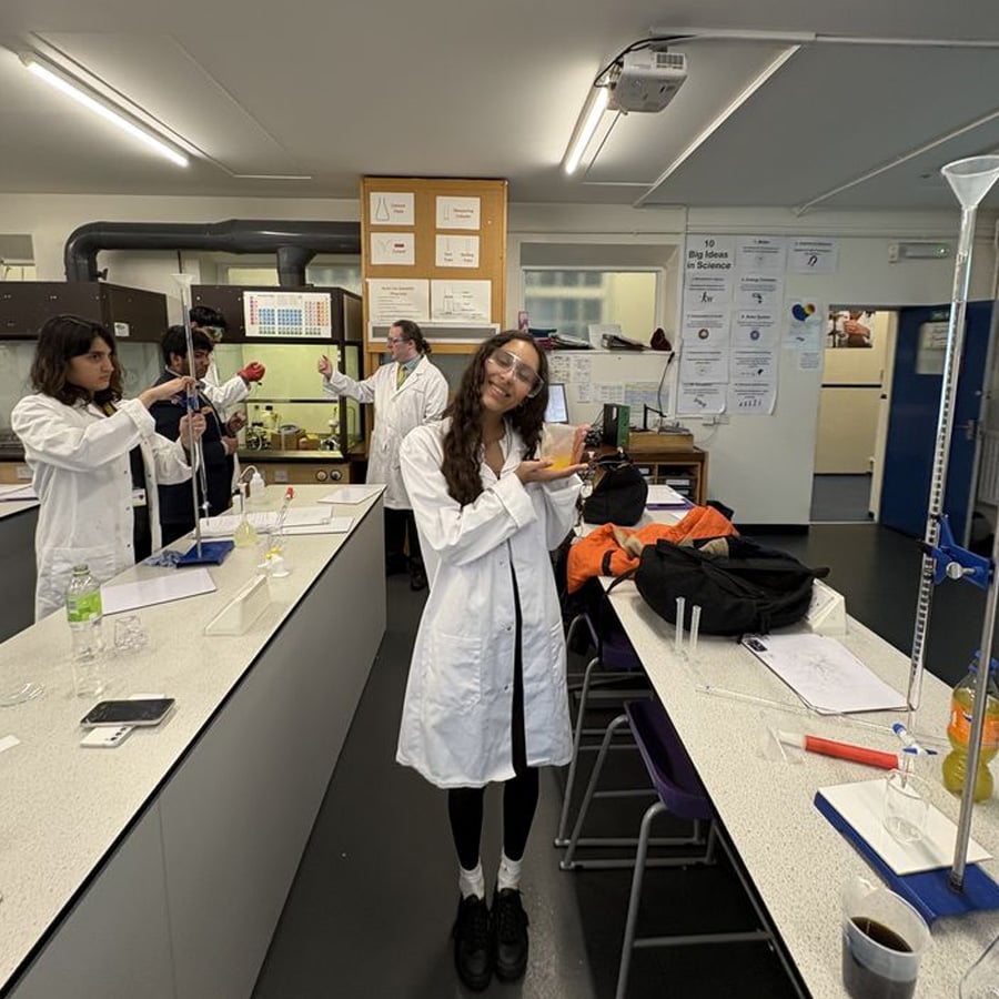 A student in a white lab coat and safety goggles poses and smiles for the camera in a science lab while classmates perform experiments in the background.