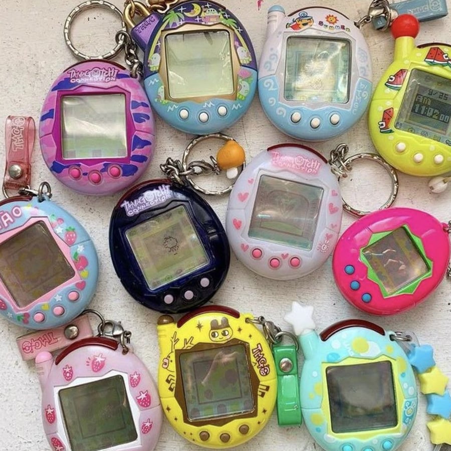 A collection of ten colorful Tamagotchi virtual pets and similar digital pets are arranged on a white surface, each with unique designs and attached keychains.