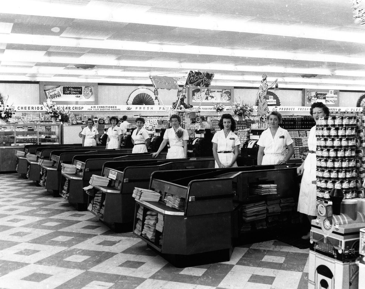 Black and white photo of six female cashiers in uniform standing at checkout lanes in a vintage grocery store, with shelves of canned goods and a “FRESH PRODUCE” sign visible in the background.