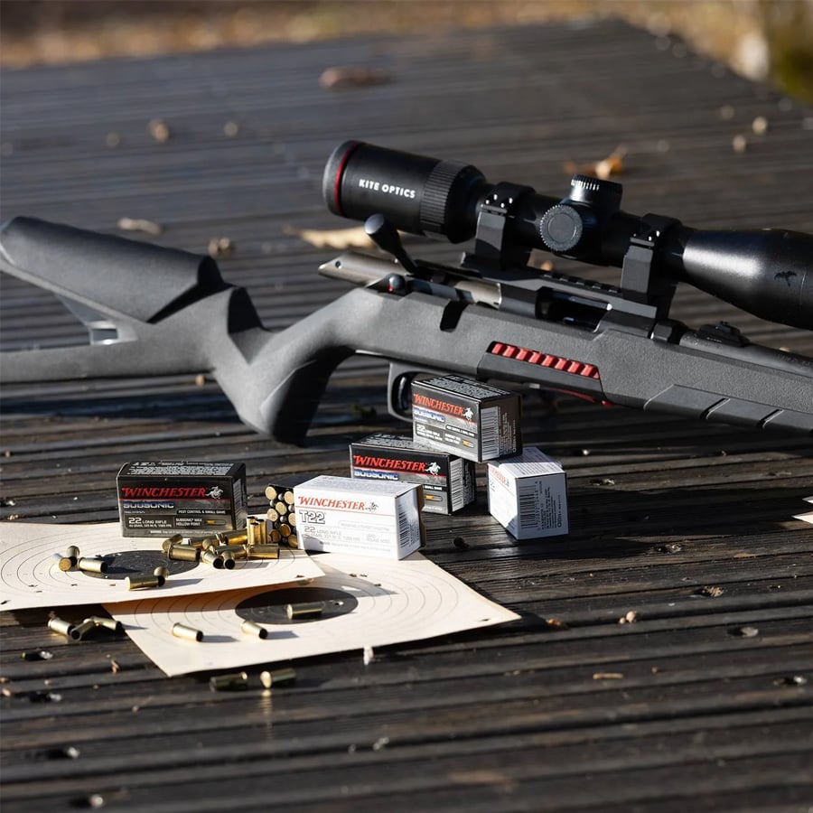 A black rifle with a scope rests on a wooden surface next to boxes of Winchester ammunition, loose bullets, and paper shooting targets.