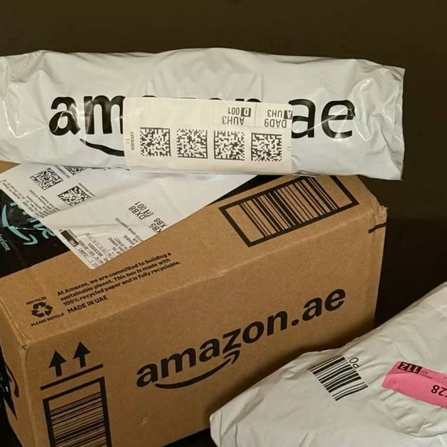 Three Amazon.ae packages are shown: one brown cardboard box and two white plastic parcels with shipping labels on them, all arranged against a plain dark background.