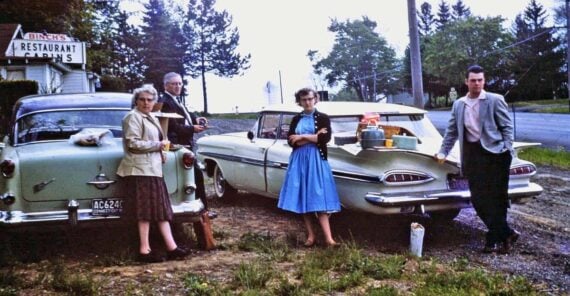 Four people stand beside two vintage cars parked on grass near a roadside diner. A picnic is set up on one car&rsquo;s trunk. The group is casually dressed, and trees and a road are visible in the background.